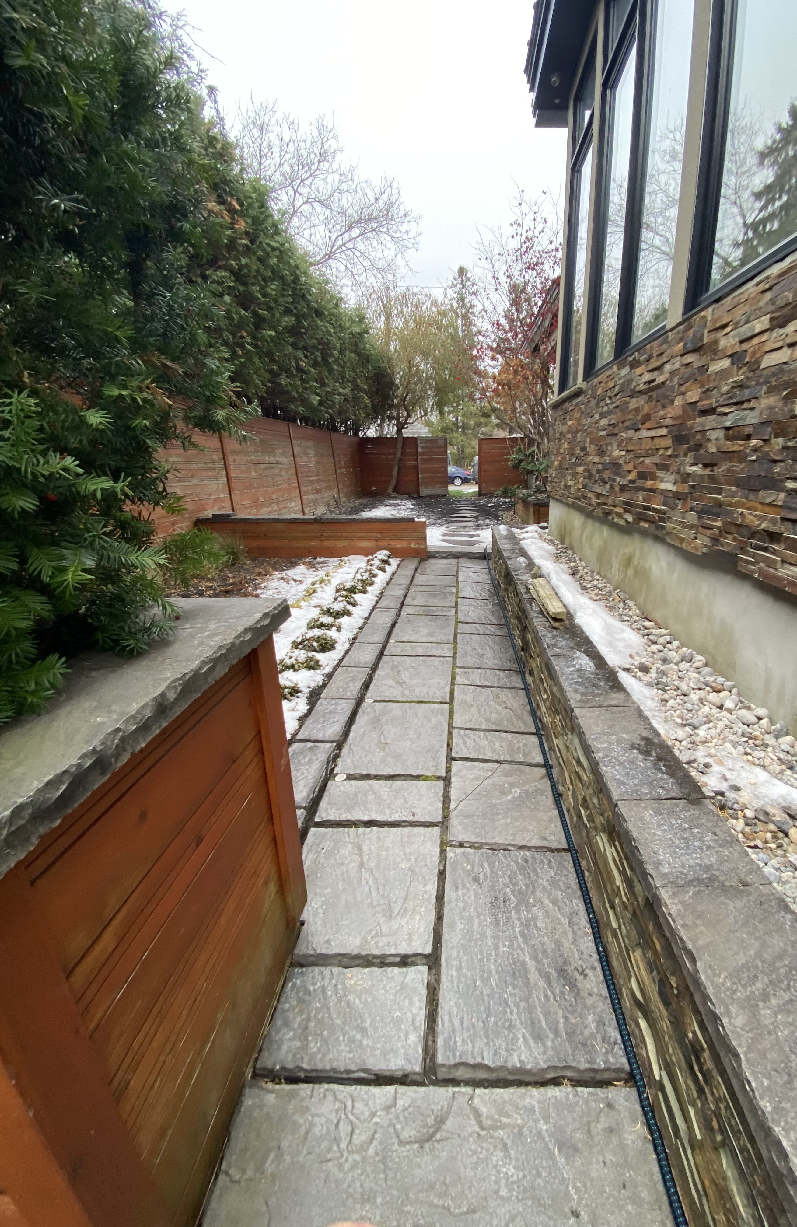Narrow stone paver walkway between a modern house and a tall evergreen hedge with light snow.